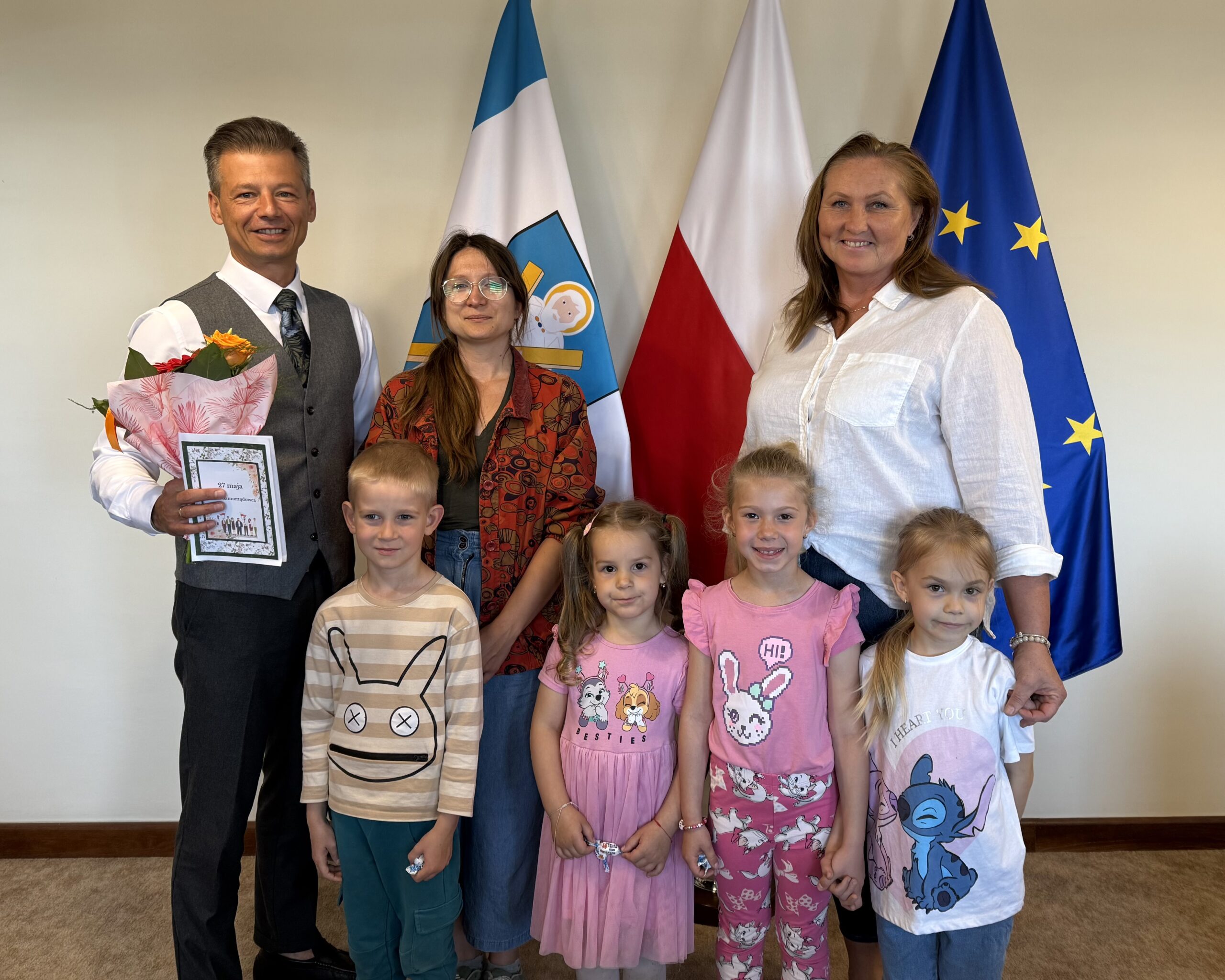 Wspólne zdjęcie zastępcy wójta z czwórką przedszkolaków i dyrektorką przedszkola. Zastępca trzyma w ręce kwiaty i laurkę.