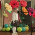 Na zdjęciu dziewczynka podczas swojego wystąpienia. W tle wystrój sceny - kolorowe duże kwiaty i balony.