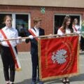 Na fotografii widoczna jest trójka uczniów: dwie dziewczyny oraz chłopak stojący pośrodku. Uczniowie trzymają nowy sztandar wykonany w kolorze czerwonym. Centralne miejsce na sztandarze zajmuje wizerunek białego orła ze złotą koroną na głowie. Powyżej umieszczono napis: „Szkoła Podstawowa im. Bohaterów Ziemi Tuchorskiej w Tuchorzy”. W tle dostrzec można innych uczniów trzymających sztandar swojej szkoły.