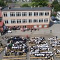 Zdjęcie wykonano z drona. Przedstawia budynek szkoły i ludzi stojących na placu przed szkołą oraz siedzących na ławkach.