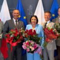 Na zdjęciu wójt, zastępca wójta oraz skarbnik gminy Przemęt. Wyżej wymienione osoby pozują do zdjęcia z bukietami róż. Na zdjęciu znajduje się także prezes Spółdzielni Socjalnej "Konwalia" Michał Żok oraz Prezes Przemęckiego Przedsiębiorstwa Komunalnego Józef Kwaśniewicz. Wszyscy pozują na tle ścianki "Gmina Przemęt" oraz flag: dwóch flag Polski i dwóch Europejskich.