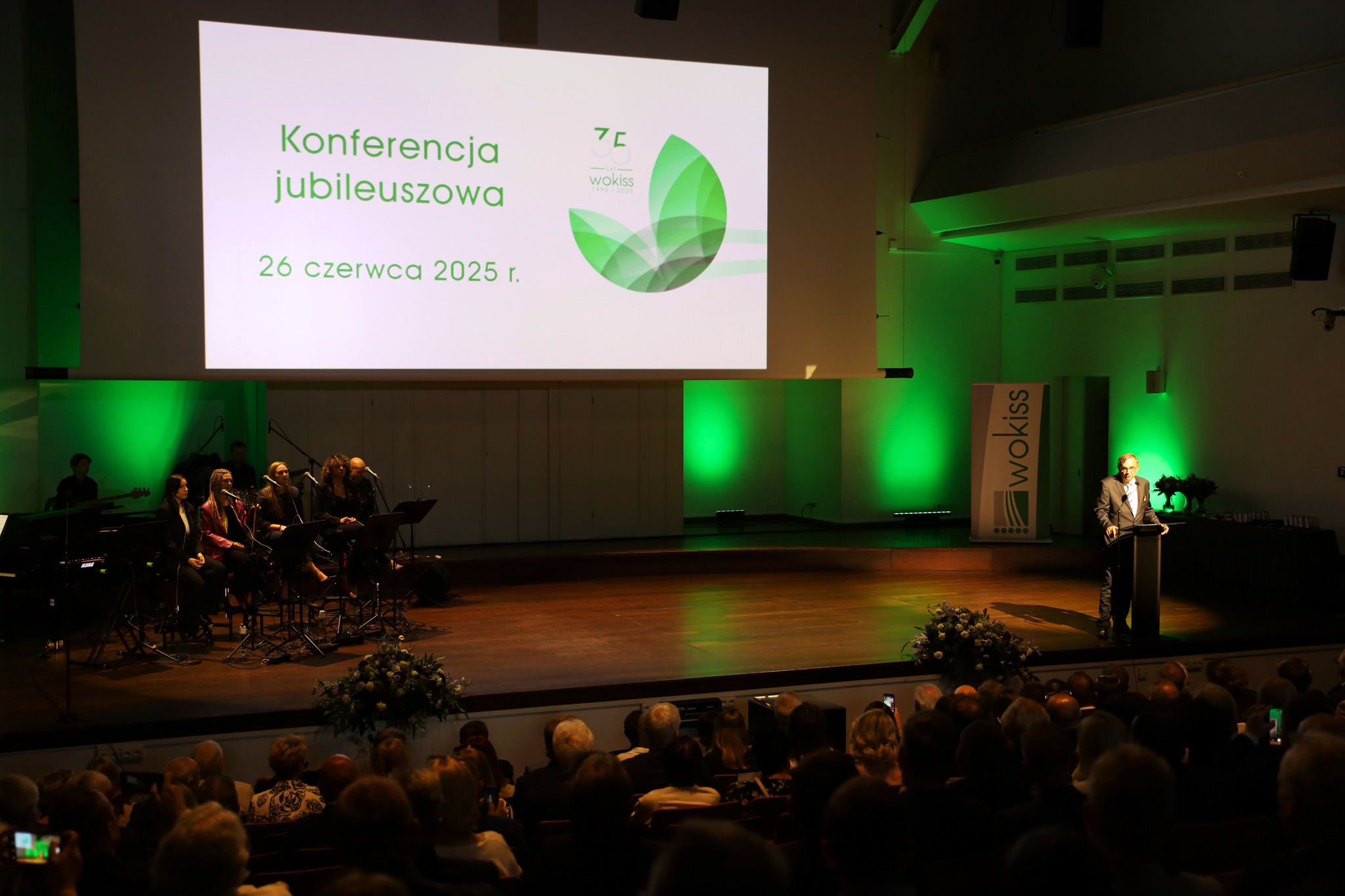 Zdjęcie przedstawia aule, na której znajdują się zaproszeni goście. Na scenie wyświetlana jest grafika przedstawiająca logo Wokissu i napis "Konferencja jubileuszowa 26 czerwca 2025 r."
