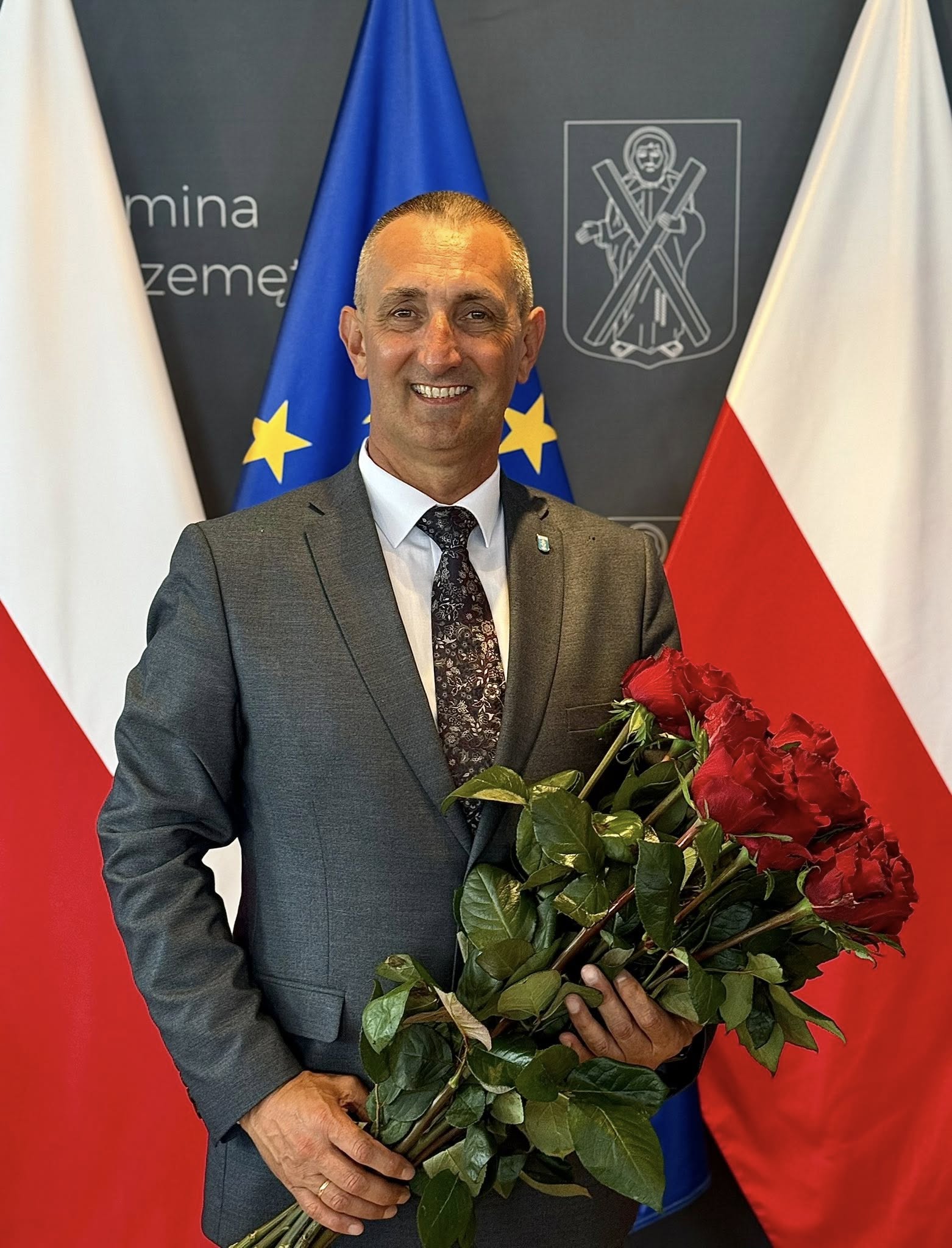 Na fotografii wójt Janusz Frąckowiak pozuje do zdjęcia z bukietem czerwonych róż. W tle ścianka gminy Przemęt oraz flagi: dwie Polski po bokach oraz jedna Europejska pośrodku.