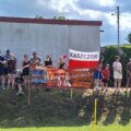 Na zdjęciu kibice. Dwie kobiety trzymają flagę Polski, na której widnieje duży napis "Kaszczor"