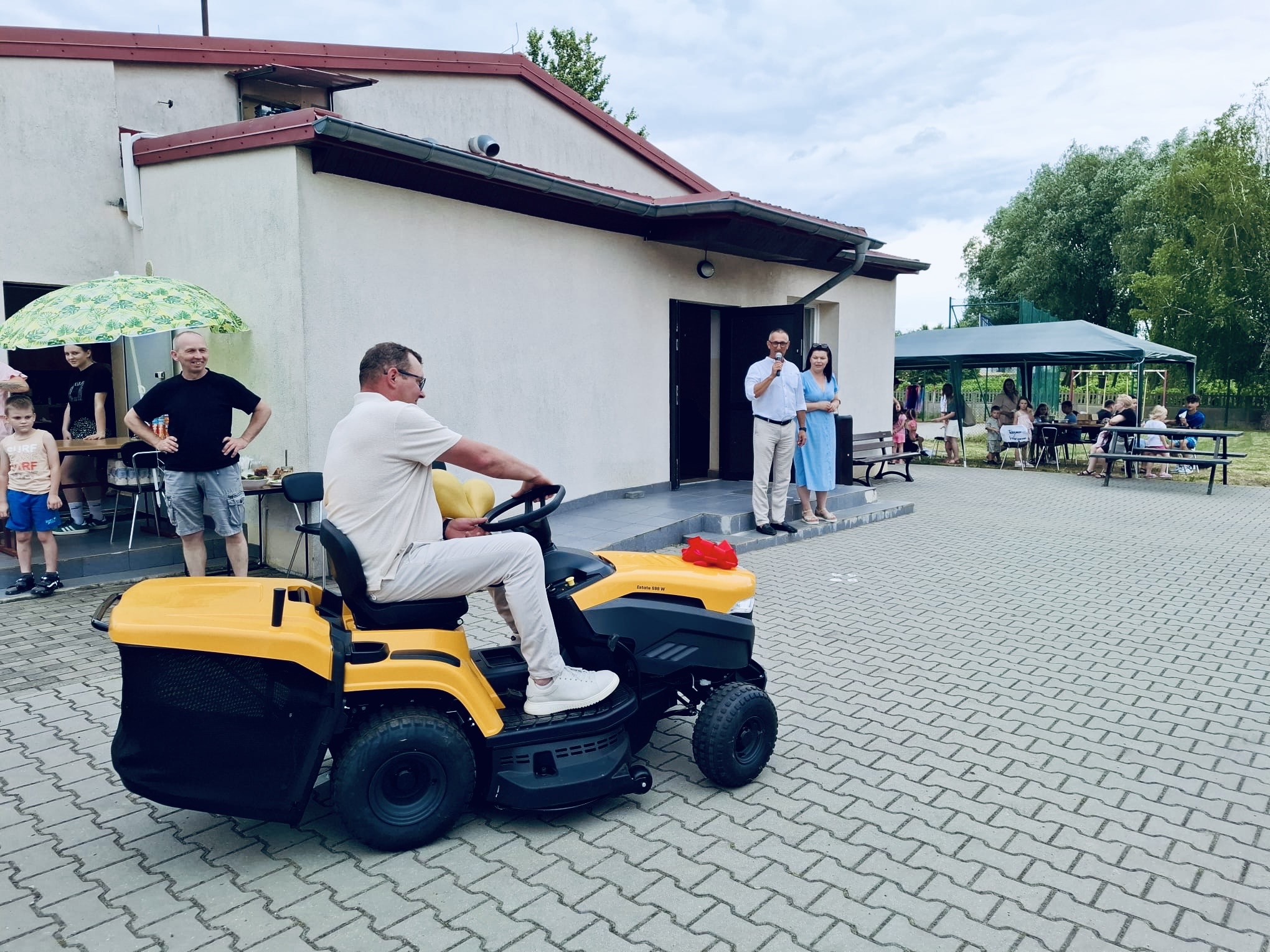 Zdjęcie przedstawia sołtysa Górska, który jedzie nowym traktorem ogrodowym. Na fotografii także wójt gminy, który przemawia przez mikrofon oraz przewodnicząca rady gminy.