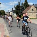 Na fotografii grupa osób biorących udział w rajdzie, jedzie rowerami ulicą. Jeden z uczestników -mężczyzna macha do kamery aparatu.