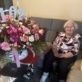 Na zdjęciu pani Stefania Łada pozuje wraz z dużym koszykiem kwiatów, do których przypięta została liczba 102.