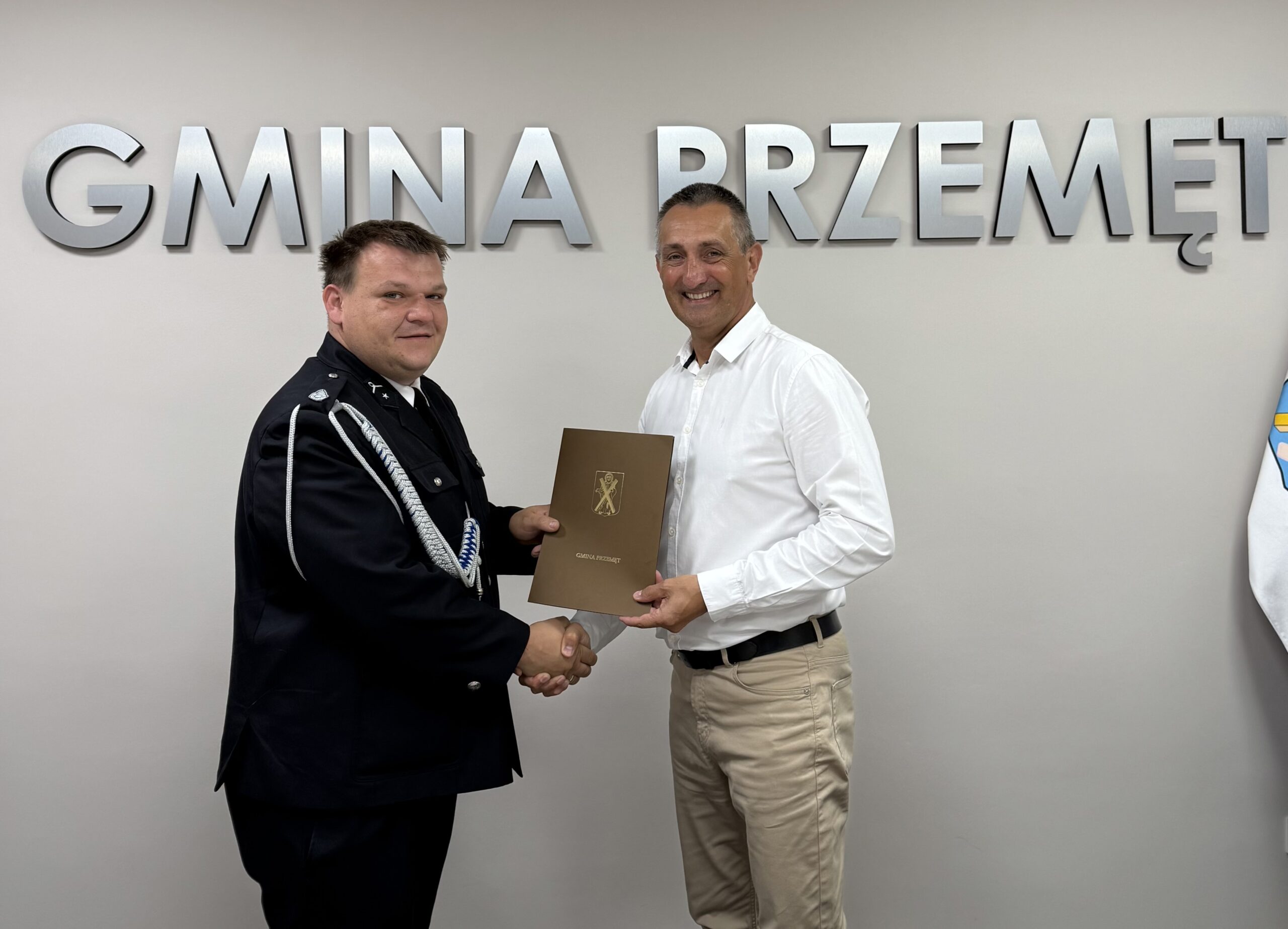 Na fotografii wójt oraz druh Robert Pawłowski pozują z umową umieszczoną w teczce. Wyżej wymienione osoby podają sobie dłoń. W tle srebrny napis: "Gmina Przemęt".