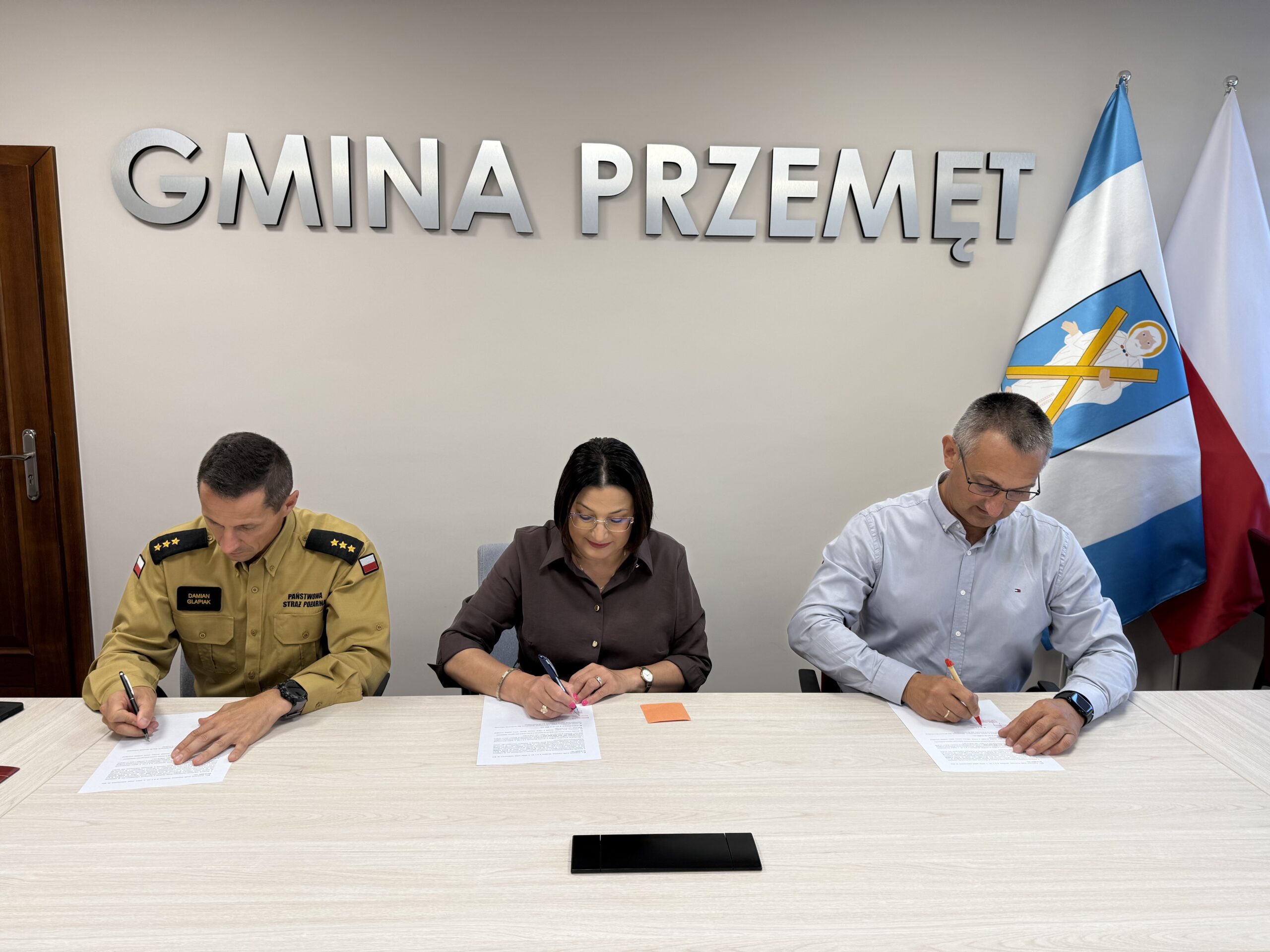 Na fotografii od prawej wójt Janusz Frąckowiak, skarbnik gminy Bożena Ćwiklińska oraz komendant powiatowy PSP w Wolsztynie – kpt. Damian Glapiak. Wszyscy podpisują umowę. W tle srebrny napis: "Gmina Przemęt" oraz flagi: Polska i z herbem gminy Przemęt.