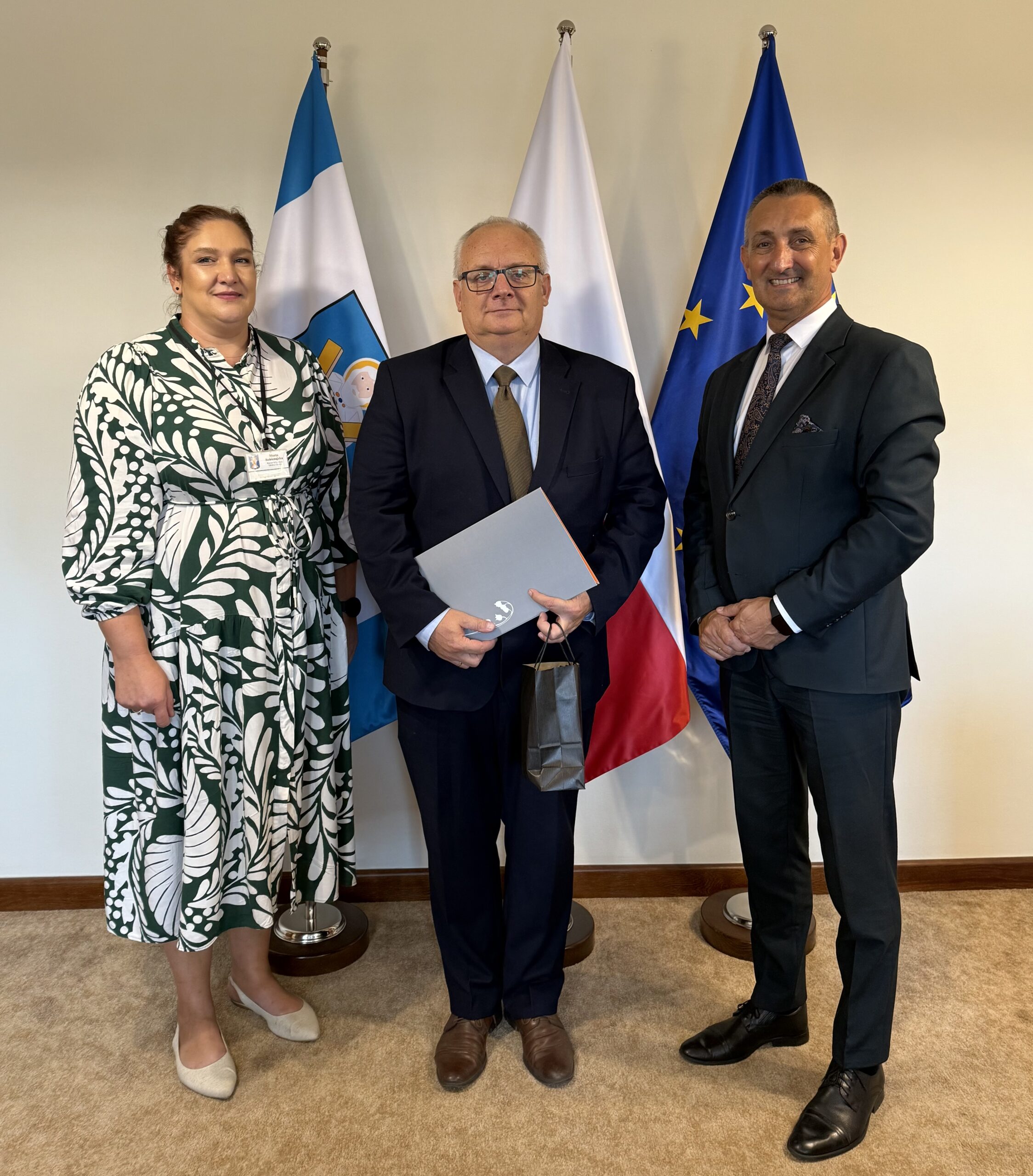 Na fotografii wójt, kierownik oświaty Marta Sobierajska oraz dyrektor Roman Smoczyk, który pozuje do zdjęcia z aktem powierzenia umieszczonym w szarej teczce oraz z upominkiem. W tle widoczne są flagi: Polski, Unii Europejskiej oraz z herbem gminy Przemęt.