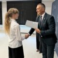 Na fotografii wójt składa gratulacje absolwentce szkoły podstawowej. W tle ścianka gminy.