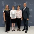 Na zdjęciu wójt, absolwentka szkoły podstawowej, która pozuje z listem gratulacyjnym oraz dyrektorka szkoły z Kaszczoru. W tle ścianka gminy.