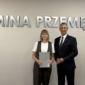 Wójt pozuje do zdjęcia z jedną z mianowanych nauczycielek. Kobieta trzyma akt mianowania w ręce. w tle srebrny napis "Gmina Przemęt" oraz flaga z herbem gminy.