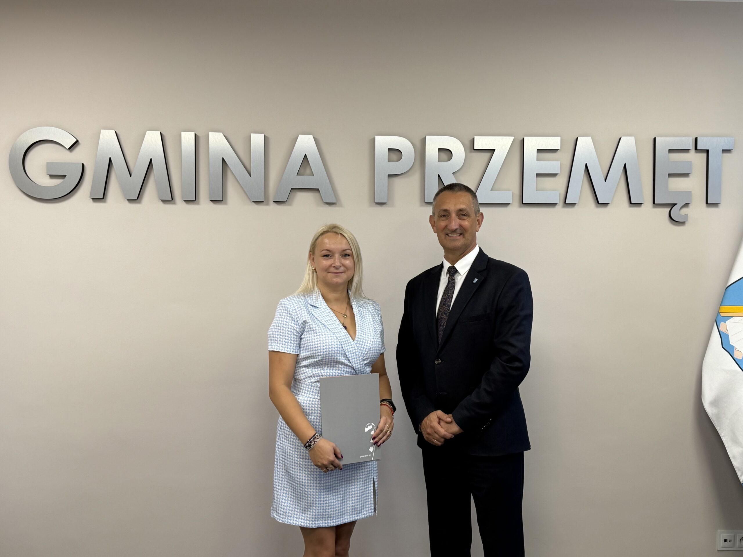 Wójt pozuje do zdjęcia z jedną z mianowanych nauczycielek. Kobieta trzyma akt mianowania w ręce. w tle srebrny napis "Gmina Przemęt" oraz flaga z herbem gminy.