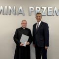 Wójt pozuje do zdjęcia z nauczycielem religii, który też jest jednocześnie księdzem. Mężczyzna trzyma akt mianowania w ręce. w tle srebrny napis "Gmina Przemęt" oraz flaga z herbem gminy.