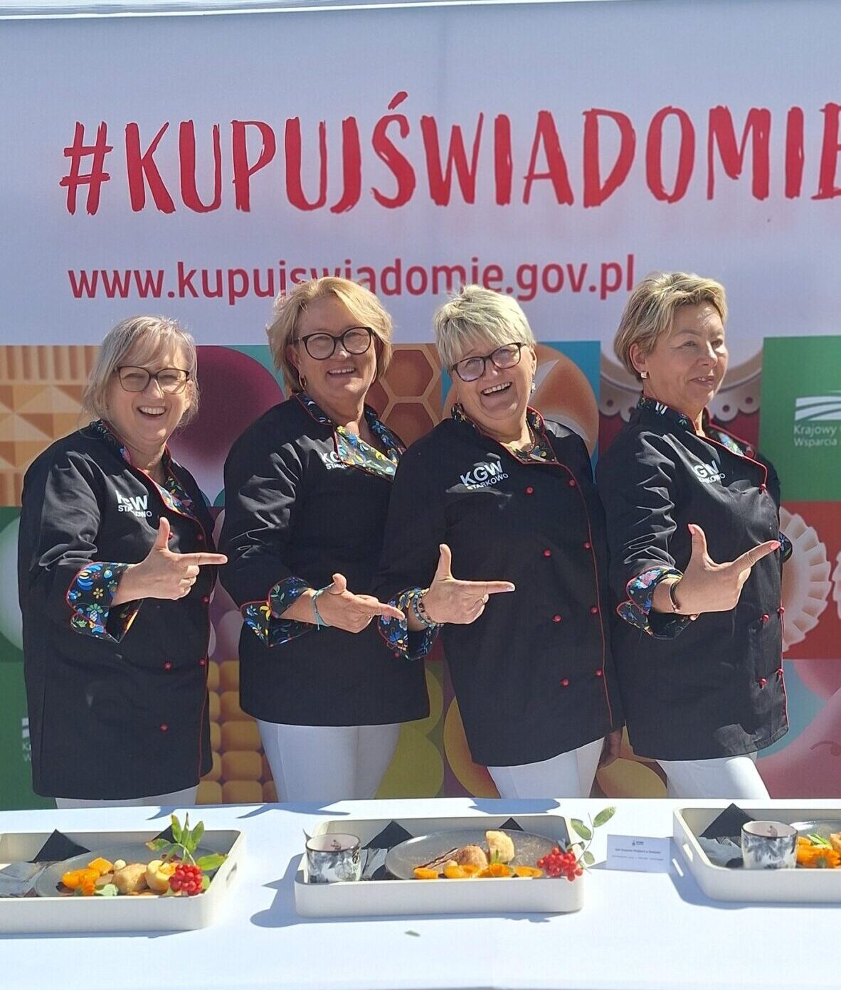 Na fotografii cztery reprezentantki KGW Starkowo pozują radośnie przed banerem akcji „Kupuj Świadomie”. Ubrane są w eleganckie czarne stroje z ludowym akcentem przy rękawach.