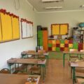 Klasyczna sala lekcyjna z zielonymi krzesłami i drewnianymi biurkami. Na biurkach leżą podręczniki, a na ścianach widoczne są dekoracje, które dodają przestrzeni ciepła. W tle stoją kolorowe szafki.