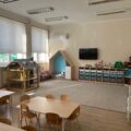 Na tym zdjęciu widzimy wnętrze przedszkola. Pomieszczenie jest jasne, przestronne, z dużymi oknami wpuszczającymi naturalne światło. Widać zabawkowy domek, który znajduje się w sali, a także różne zestawy edukacyjne i zabawki ułożone w szafkach.