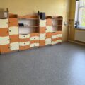 To zdjęcie przedstawia przestrzeń z meblami w klasycznych, pastelowych kolorach. Widać regały z szufladami w odcieniach pomarańczowego i kremowego. Meble są nowoczesne, z zaokrąglonymi krawędziami. Widoczna też nowa wykładzina.