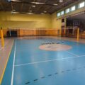 To zdjęcie przedstawia halę sportową. Widoczna jest siatka do siatkówki, kilka piłek leżących na podłodze oraz drewniane trybuny w tle.