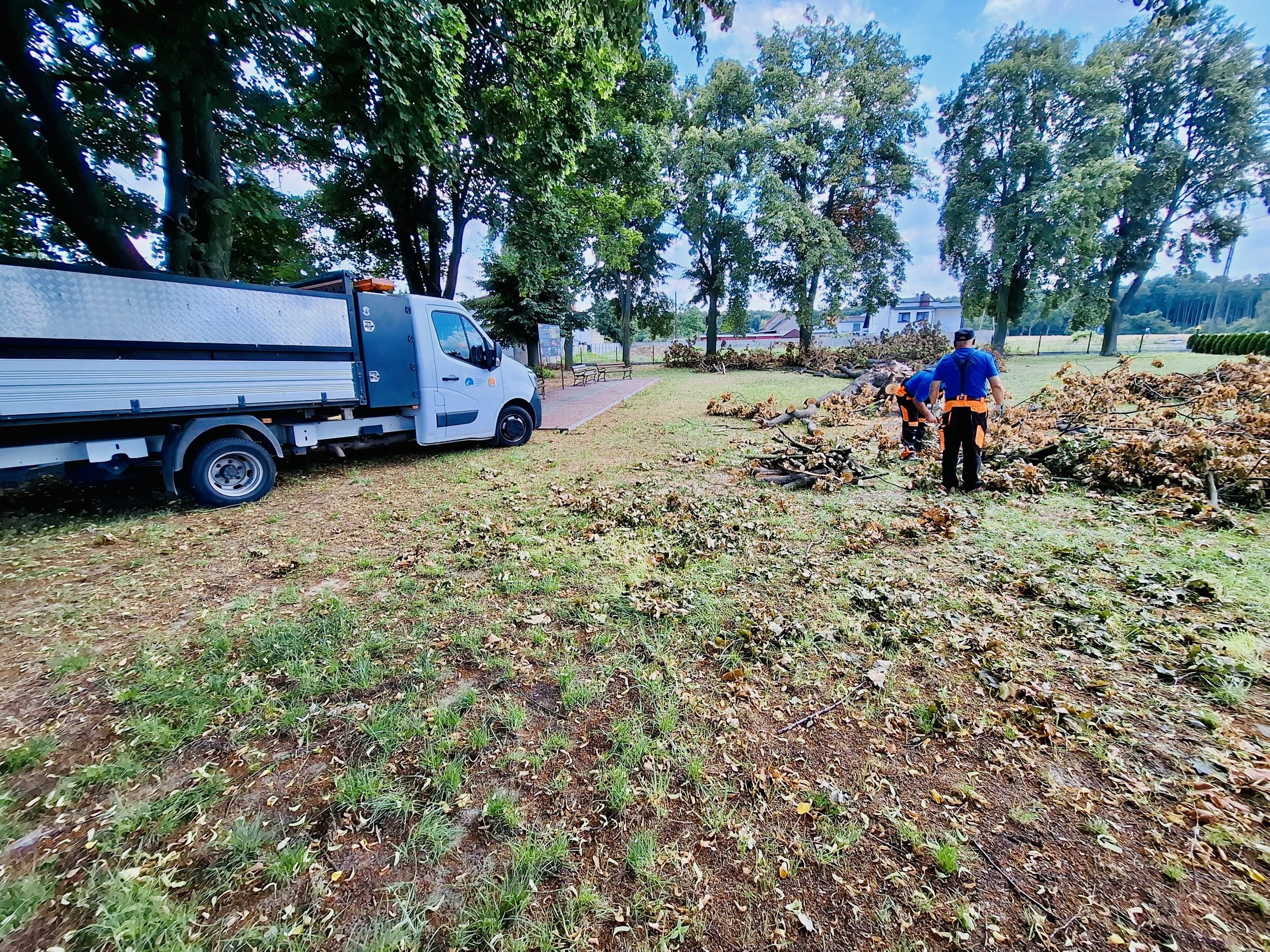 Na zdjęciu widzimy pracowników Przemęckiego Przedsiębiorstwa Komunalnego w trakcie porządkowania terenów zielonych. Dwóch mężczyzn w roboczych ubraniach usuwa połamane gałęzie i konary drzew, które leżą na trawniku. Obok nich stoi samochód służbowy, którym przewożą zebrany materiał.