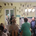 Grupa zwiedzających słucha opowieści leśnika w jednej z sal edukacyjnych. Wnętrze udekorowane jest licznymi porożami oraz trofeami myśliwskimi. Na ścianach znajdują się także edukacyjne plakaty przedstawiające chronione gatunki zwierząt.