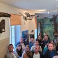 Grupa zwiedzających słucha opowieści leśnika w jednej z sal edukacyjnych. Wnętrze wypełniają sztuczne ptaki oraz inne zwierzęta.