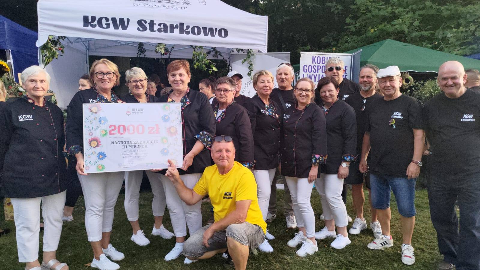Na zdjęciu znajduje się grupa osób reprezentujących Koło Gospodyń Wiejskich Starkowo oraz sołtys Starkowa (mężczyzna w żółtej koszulce z napisem "KGW Starkowo") Członkinie KGW ubrane są w jednakowe stroje – czarne koszule z kolorowymi mankietami i logiem KGW Starkowo oraz białe spodnie. Obok nich stoją mężczyźni wspierający KGW, ubrani w czarne koszulki z tym samym logo. W centrum zdjęcia trzymany jest duży symboliczny czek na kwotę 2000 zł, który grupa otrzymała jako nagrodę za zajęcie III miejsca w konkursie kulinarnym "Bitwa Regionów".