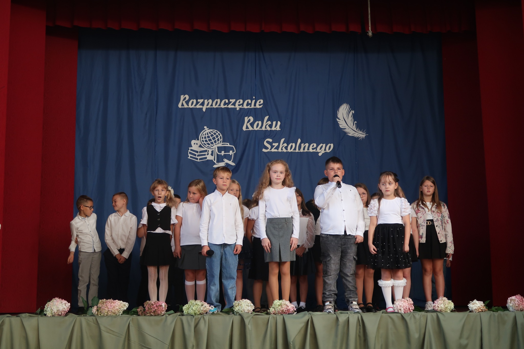 Na zdjęciu znajduje się grupa uczniów występujących na scenie, w ramach części artystycznej inauguracji roku szkolnego. Wszyscy ubrani są w galowe stroje. Scena została udekorowana niebieską kotarą z napisem „Rozpoczęcie Roku Szkolnego”.
