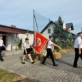 Na zdjęciu trwa przemarsz pocztu sztandarowego. Mężczyźni i kobieta ubrani w galowe mundury niosą czerwone sztandary z polskimi symbolami.