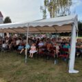 Na zdjęciu widzimy tłum ludzi siedzących pod dużym białym namiotem na świeżym powietrzu. Są tam osoby w różnym wieku – dzieci, dorośli i starsi. W tle znajduje się duża, zielona dmuchana zjeżdżalnia dla dzieci.