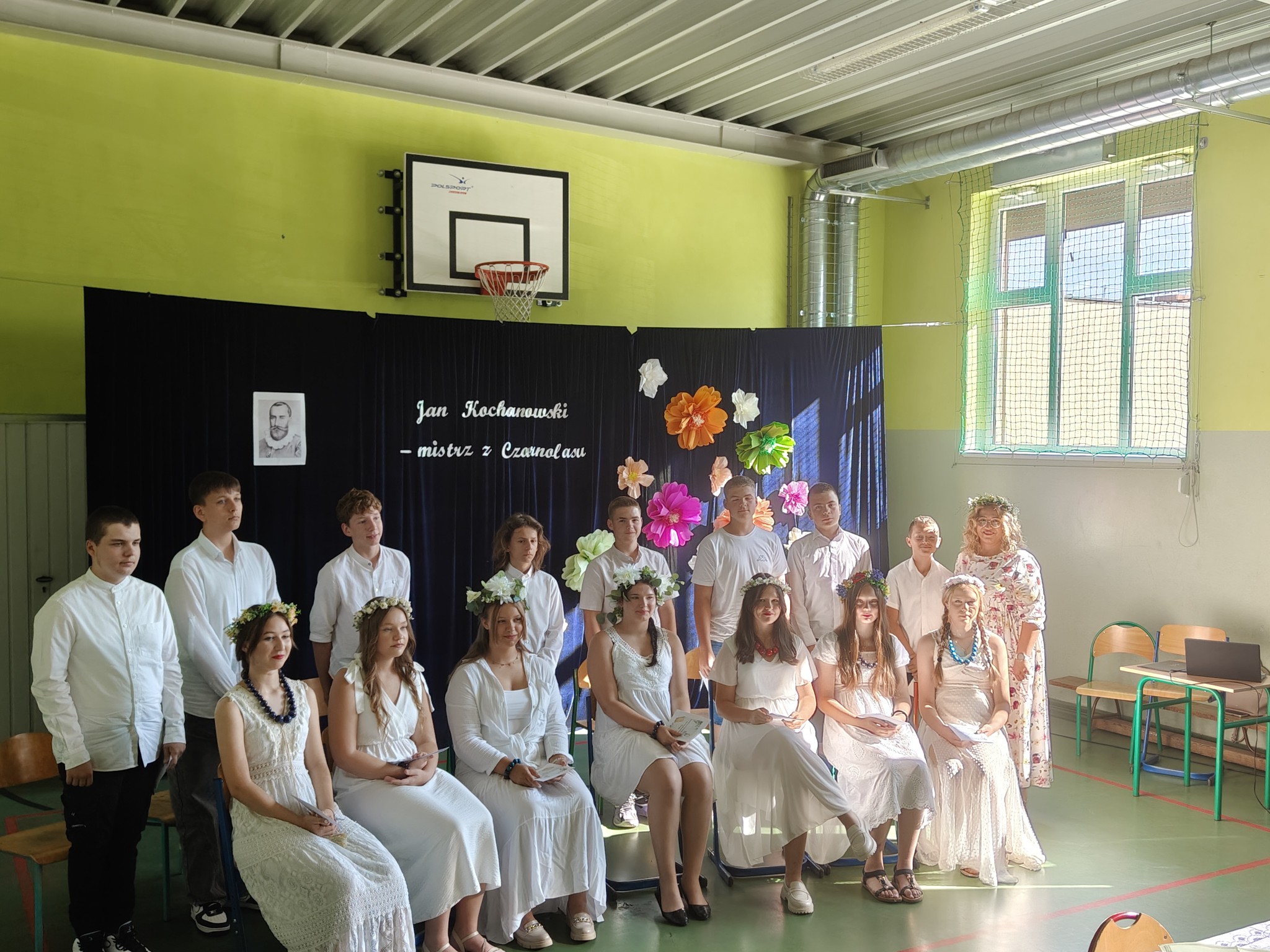 Na zdjęciu grupa uczniów, którzy pozują do wspólnego zdjęcia. Większość z nich ma na głowach wianki, a wszyscy są ubrani na biało. Uczniowie są ustawieni w rzędach – niektórzy siedzą, inni stoją. Tło stanowi granatowa kurtyna, która jest udekorowana papierowymi kwiatami oraz widnieje na niej napis "Jan Kochanowski - mistrz z Czarnolasu".