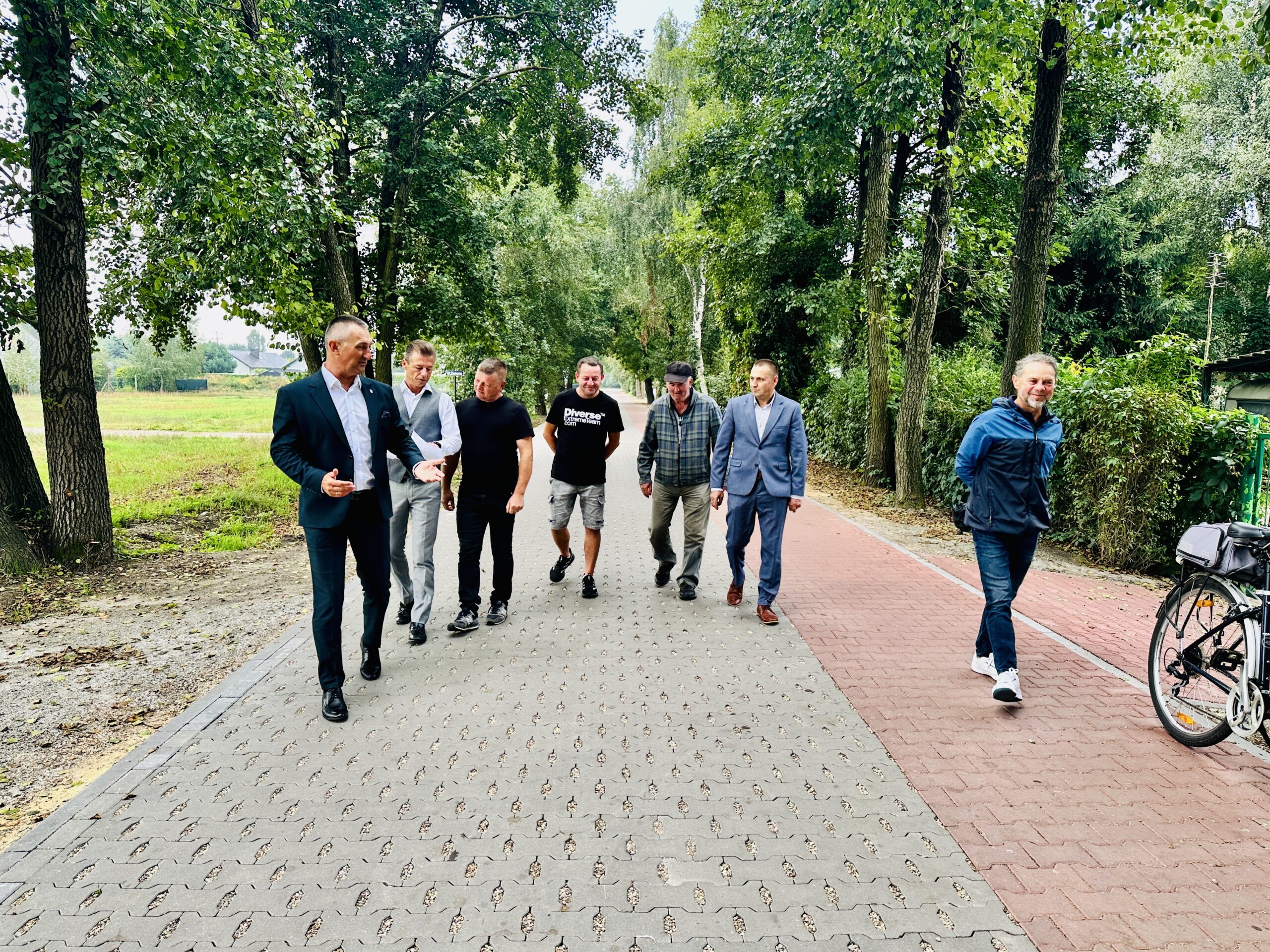 Na fotografii widać grupę osób uczestniczących w odbiorze inwestycji, idących wzdłuż nowo wybudowanej drogi. Po obu stronach trasy rosną rzędy drzew.