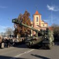 Na zdjęciu widoczny czołg wojskowy, który jedzie drogą. Na zdjęciu widoczni także ludzie.