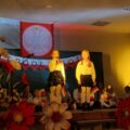 Na zdjęciu dwie dziewczyny podczas wystąpienia na scenie. Scena udekorowana jest w duże, sztuczne kwiaty koloru białego i czerwonego oraz widoczny jest napis "niepodległość". W tle widać inne osoby. Wszyscy trzymają w rękach małe flagi Polski na patyku.