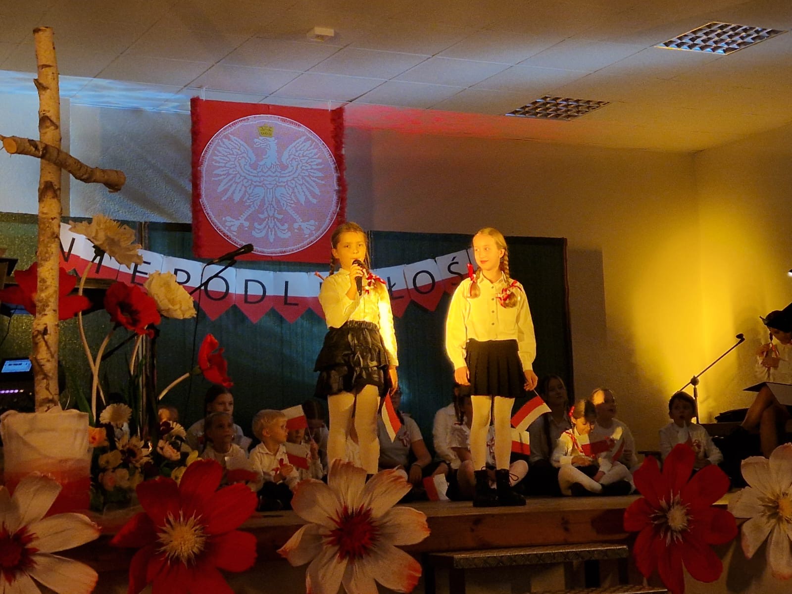 Na zdjęciu dwie dziewczyny podczas wystąpienia na scenie. Scena udekorowana jest w duże, sztuczne kwiaty koloru białego i czerwonego oraz widoczny jest napis "niepodległość". W tle widać inne osoby. Wszyscy trzymają w rękach małe flagi Polski na patyku.