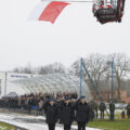 Na zdjęciu strażacy składają salut pod unoszącą się na podnośniku flagą Polski. W tle zgromadzona społeczność lokalna.