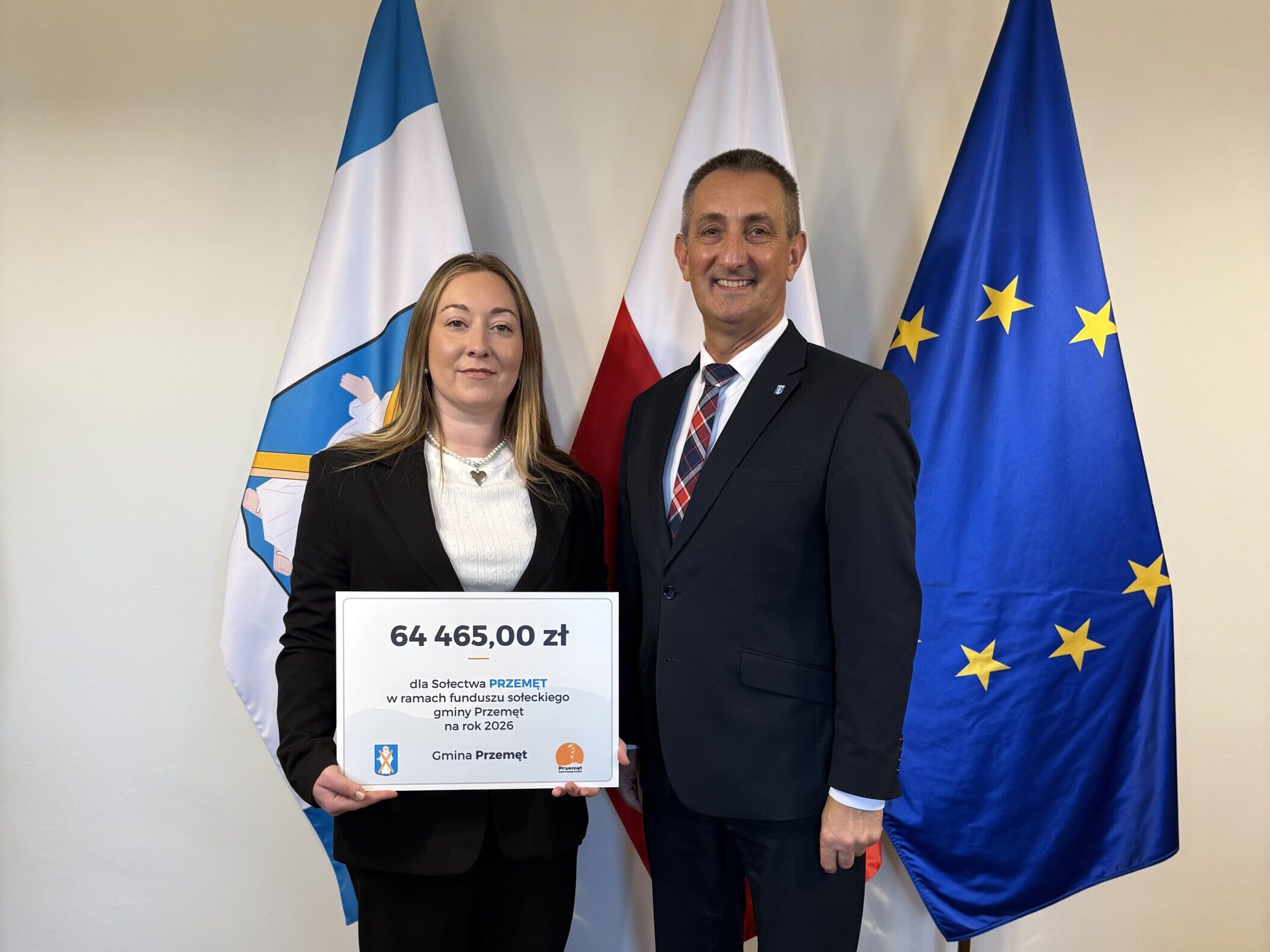 Na fotografii wójt wraz z sołtysem Przemętu pozują z czekiem. W tle widoczne są flagi: Polski, Unii Europejskiej oraz z herbem gminy Przemęt.