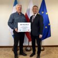 Na fotografii wójt wraz z sołtysem Solca pozują z czekiem. W tle widoczne są flagi: Polski, Unii Europejskiej oraz z herbem gminy Przemęt.