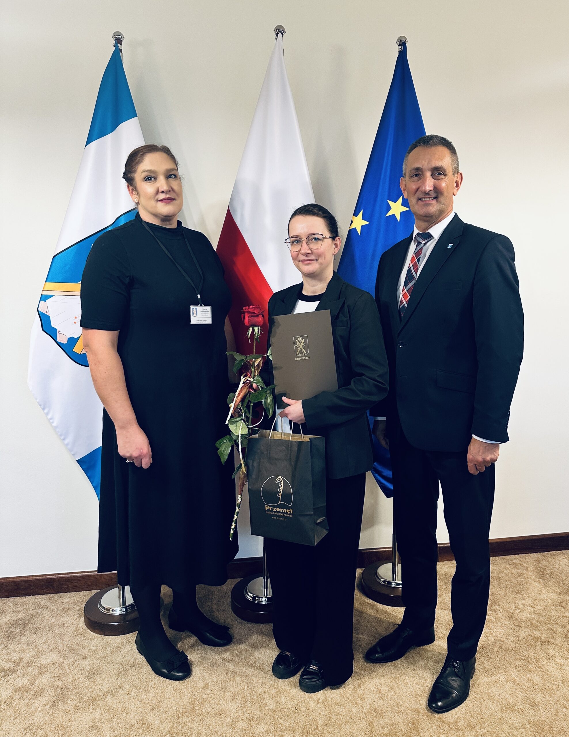 Na zdjęciu wójt, kierownik wydziału oświaty oraz pani Anna Rynek, która pozuje z aktem mianowania, różą i upominkiem.