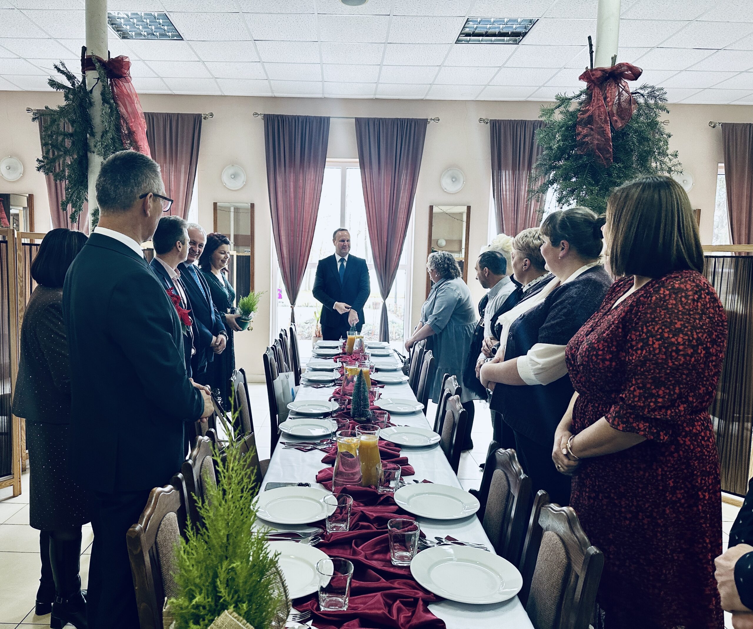 Na fotografii widoczne osoby będące na wigilii. Wszyscy stoją przy dużym stole i słuchają przemowy prezesa konwalii.