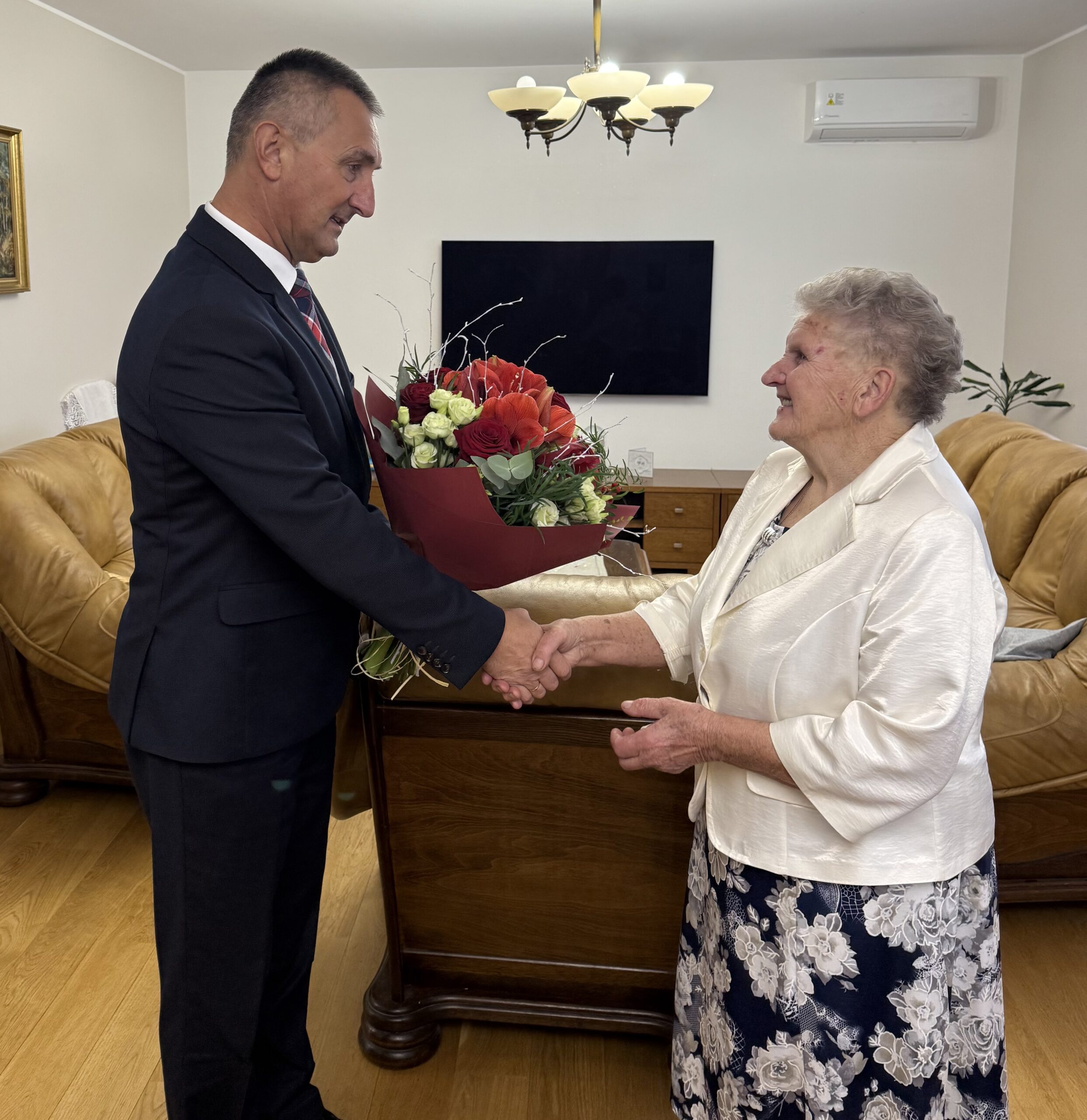Na zdjęciu wójt wręcza jubilatce bukiet kwiatów i składa życzenia.