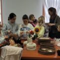 Dzieci stoją przy stole pełnym włóczek i rękodzieła, ucząc się szydełkowania i robótek ręcznych. Na stole znajdują się wykonane koszyki, czapki oraz inne wyroby z włóczki.