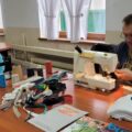 Przy stole siedzi mężczyzna obsługujący maszynę do szycia, a obok niego kobieta i dziewczynka przyglądają się pracy. Na blacie znajdują się nici, materiały i akcesoria krawieckie.