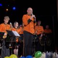Dyrygent przemawia przez mikrofon, w tle orkiestra.